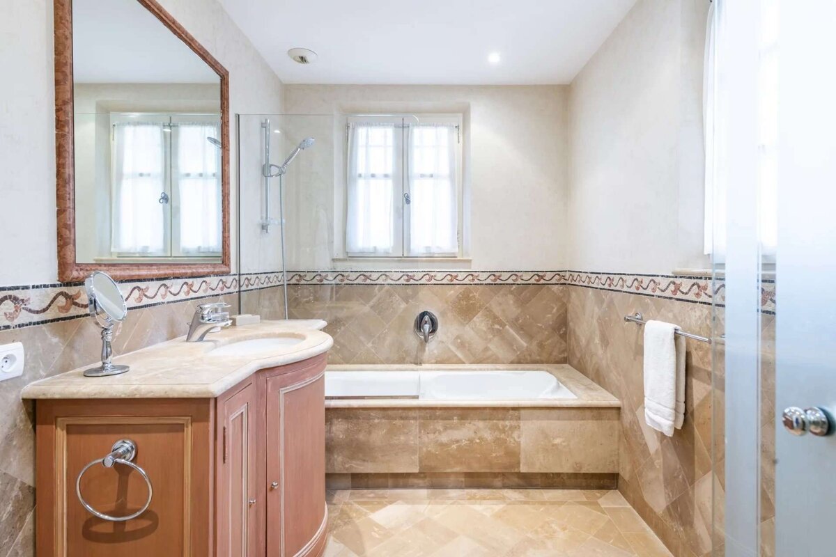 A classically designed bathroom features light-toned marble tiles, a built-in bathtub with shower, wooden vanity, sink, mirror, and window. Decorative border tiles add detail.