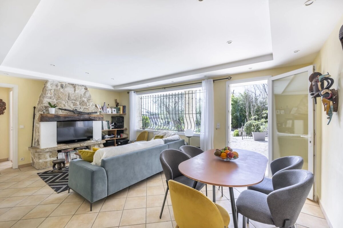 A sunny living/dining room with yellow walls, a stone fireplace, blue sofa, and a dining table with grey & yellow chairs, opening to a garden.