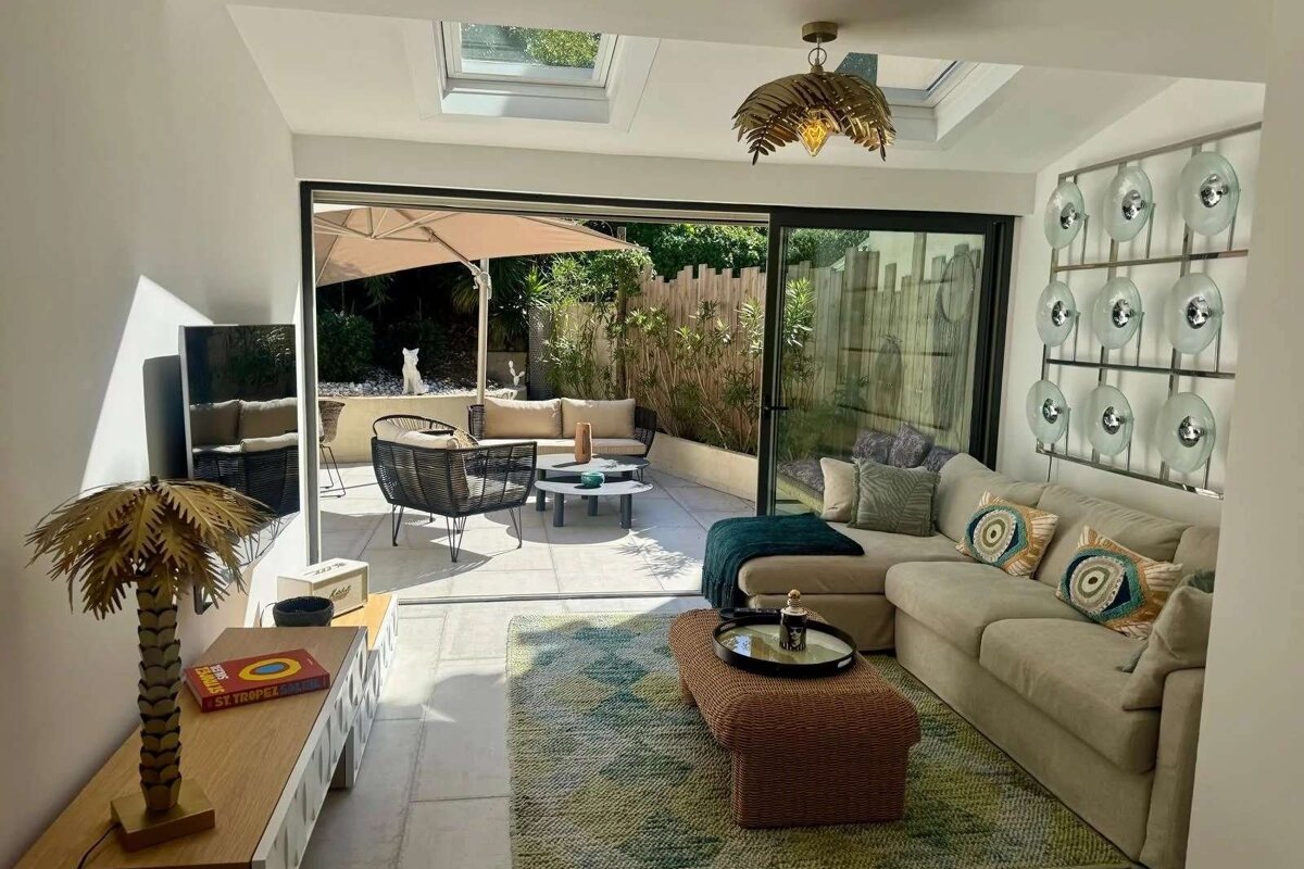 Bright, modern indoor-outdoor living space with large sliding doors leading to a sunny patio. Features stylish furniture, palm decor, and skylights.