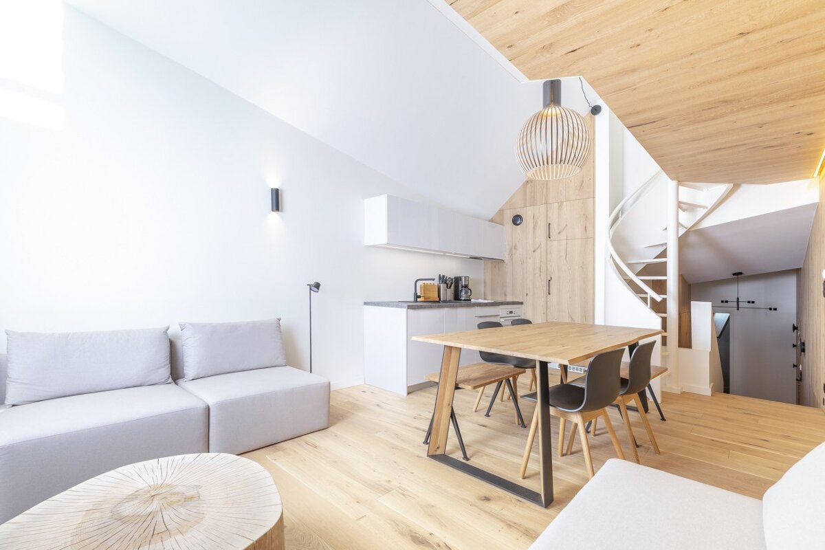 Bright modern apartment with light wood flooring, a grey sofa, a dining table, a kitchen, and a spiral staircase.
