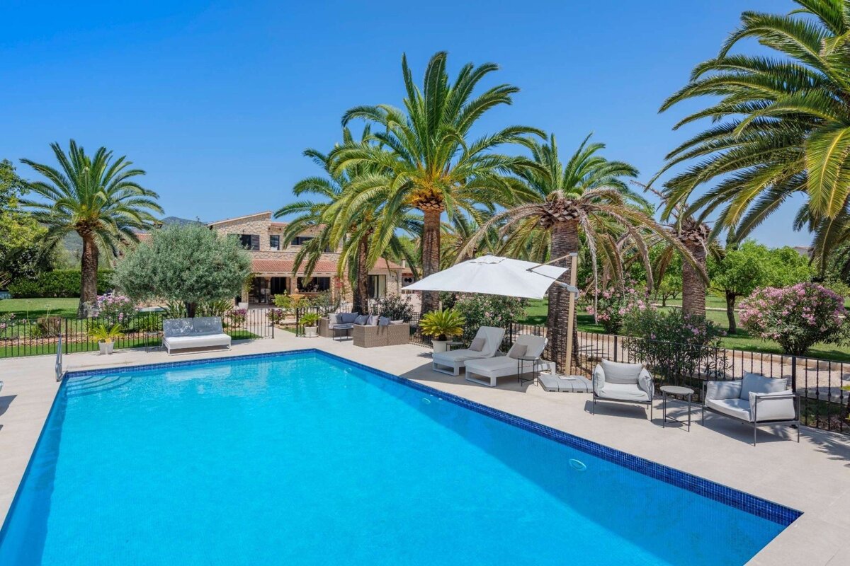 A bright blue swimming pool with lounge chairs and palm trees, set against a large building under a clear, sunny sky.