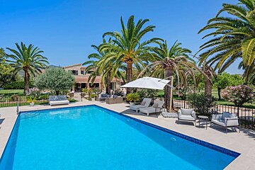 A clear blue swimming pool bordered by palm trees, outdoor seating, and a large house, all under a bright sunny sky.