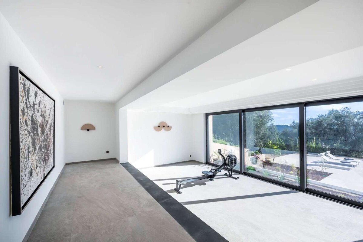 A bright, modern home gym featuring a rowing machine, large windows with a scenic outdoor view, white walls, and abstract art.