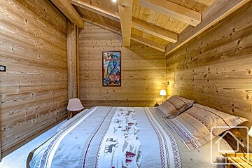 Cozy, rustic wooden bedroom with a large bed featuring ski-themed bedding, a vibrant abstract painting, and a small pink chair.