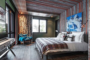 Modern rustic bedroom with a plaid bed, wood floor, striped walls & ceiling, large window, blue armchair, and mountain art.