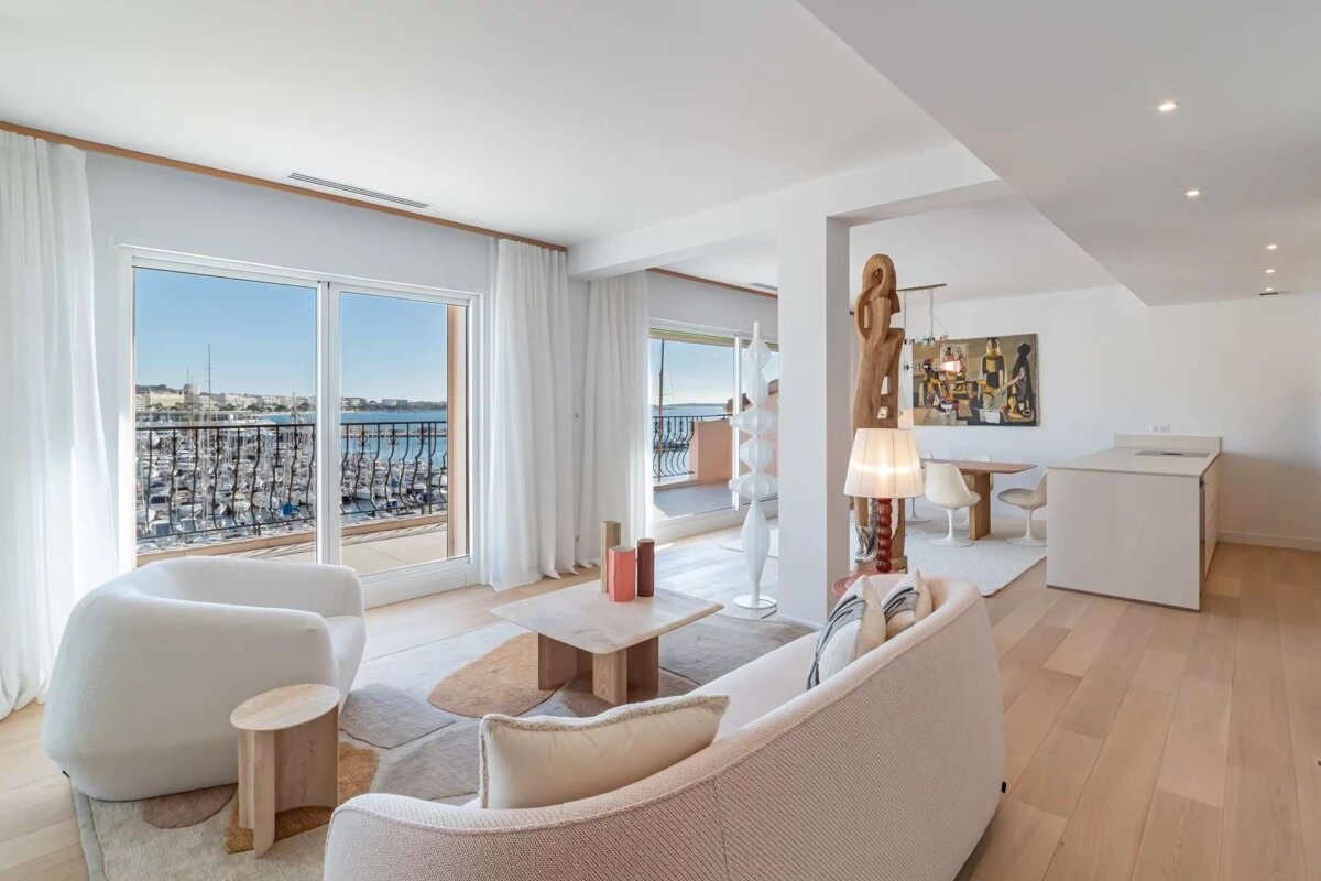 A bright, modern living room with stylish, light-colored furniture and a balcony overlooking a bustling marina and the sea.