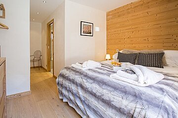 Cozy bedroom with a wooden panel accent wall, a bed with a grey textured blanket and folded robes, and light wood floors.