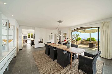 Modern dining room with a long wooden table and black chairs, leading to a sunny outdoor patio with an ocean view and stone wall.