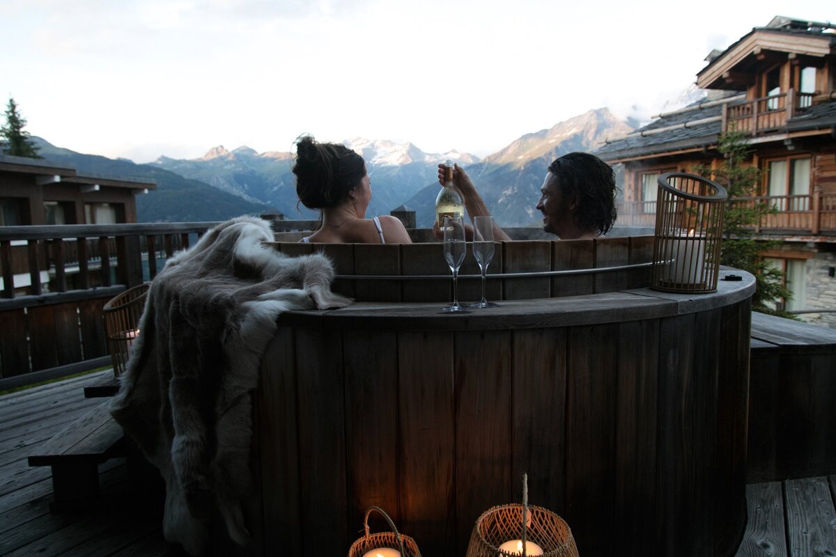 A couple relaxes in a wooden hot tub on a mountain chalet balcony, sharing champagne with a stunning alpine view at dusk.