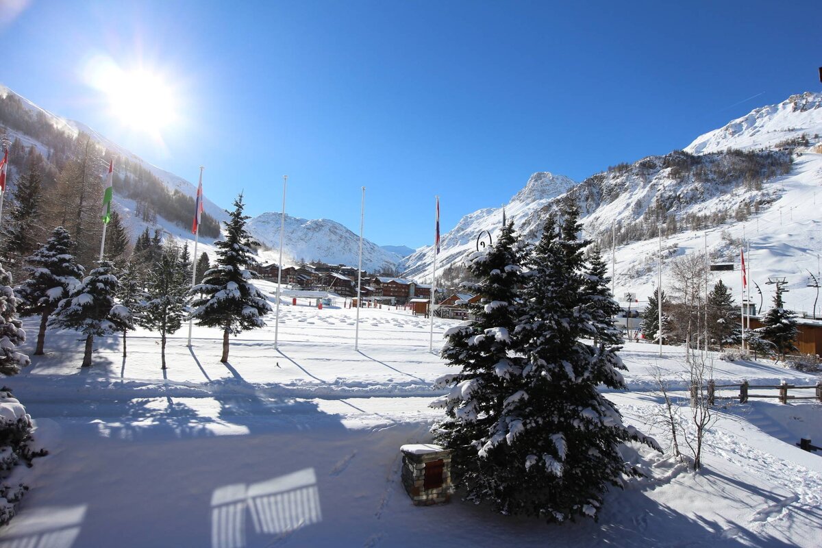 A bright, sunny winter day in a snow-covered mountain valley. Pine trees line the snowy ground with flags, and a ski resort nestles in the distance.