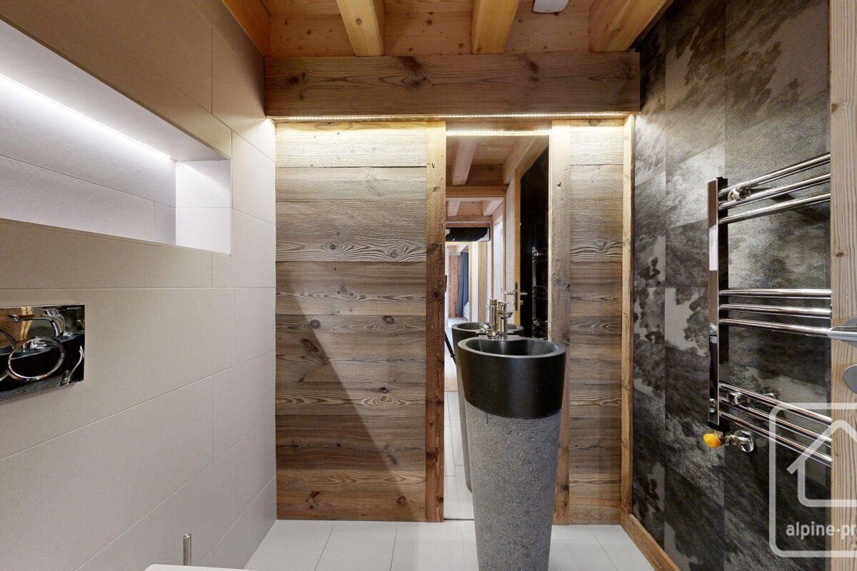 A modern rustic bathroom featuring a unique stone pedestal sink, warm wooden paneling, white tiled walls with LED lighting, and a dark patterned accent wall.