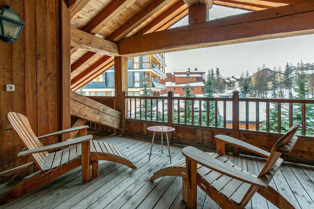 Rustic wooden balcony of a ski chalet with two lounge chairs overlooking a snowy village.