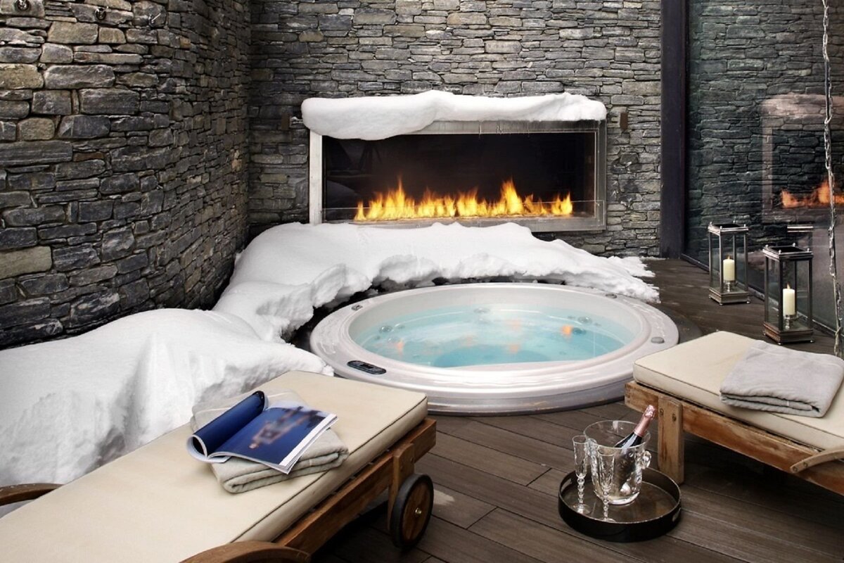 A luxurious outdoor hot tub surrounded by snowdrifts, a stone wall, and a long modern fireplace, with lounge chairs and champagne nearby.