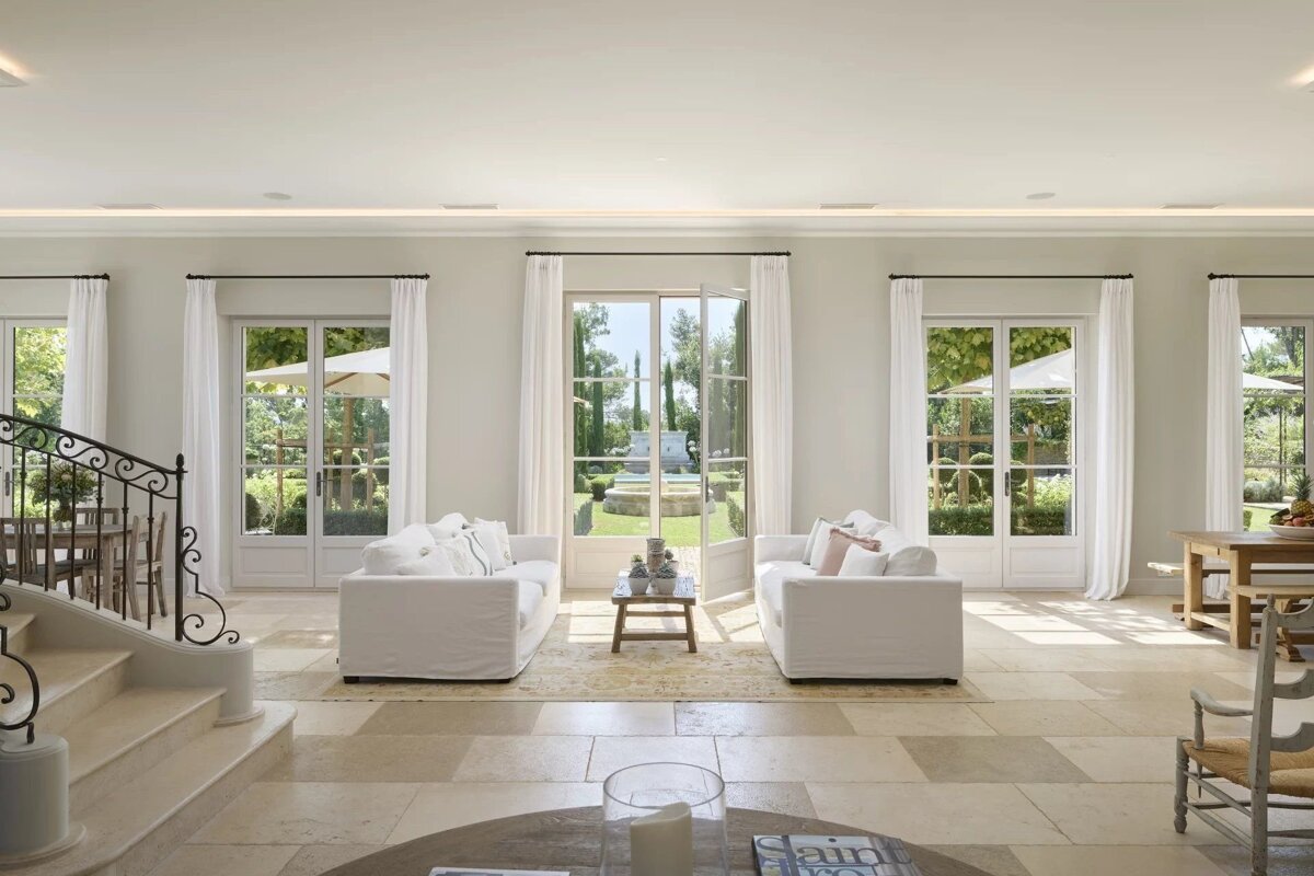 Elegant living room with white sofas, French doors leading to a garden, and a grand staircase.