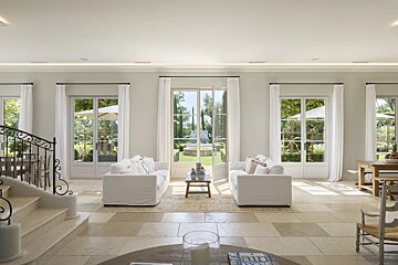 Elegant living room with white sofas, French doors leading to a garden, and a grand staircase.
