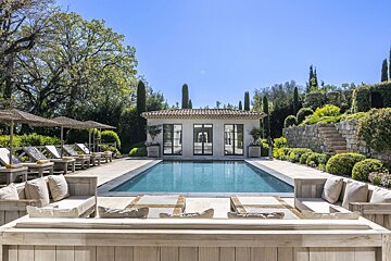 A luxurious outdoor swimming pool surrounded by lounge seating, sunbeds, and a stone pool house in a lush, sunny garden.