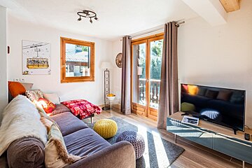 A cozy, bright living room features a sofa with colorful pillows, a TV, wooden-framed window and balcony doors, and sunlit floor.