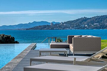 Luxurious infinity pool deck with modern furniture overlooks a vast blue sea and distant mountains under a clear sky.