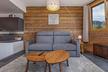 A cozy, modern chalet-style living space featuring a gray sofa, wood-paneled walls, a kitchenette, and a window overlooking mountains.
