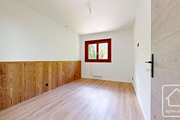 Bright, empty room with light wood flooring, white walls, and natural wood paneling below. A red-framed window reveals a green outdoor view.