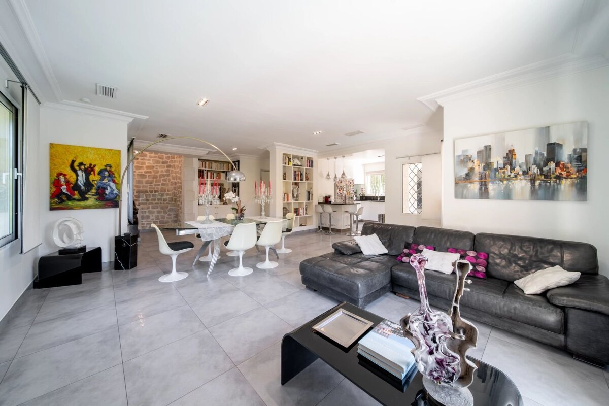 Modern open-plan living room featuring a dark leather sectional, white dining set, tiled floors, and vibrant artworks.