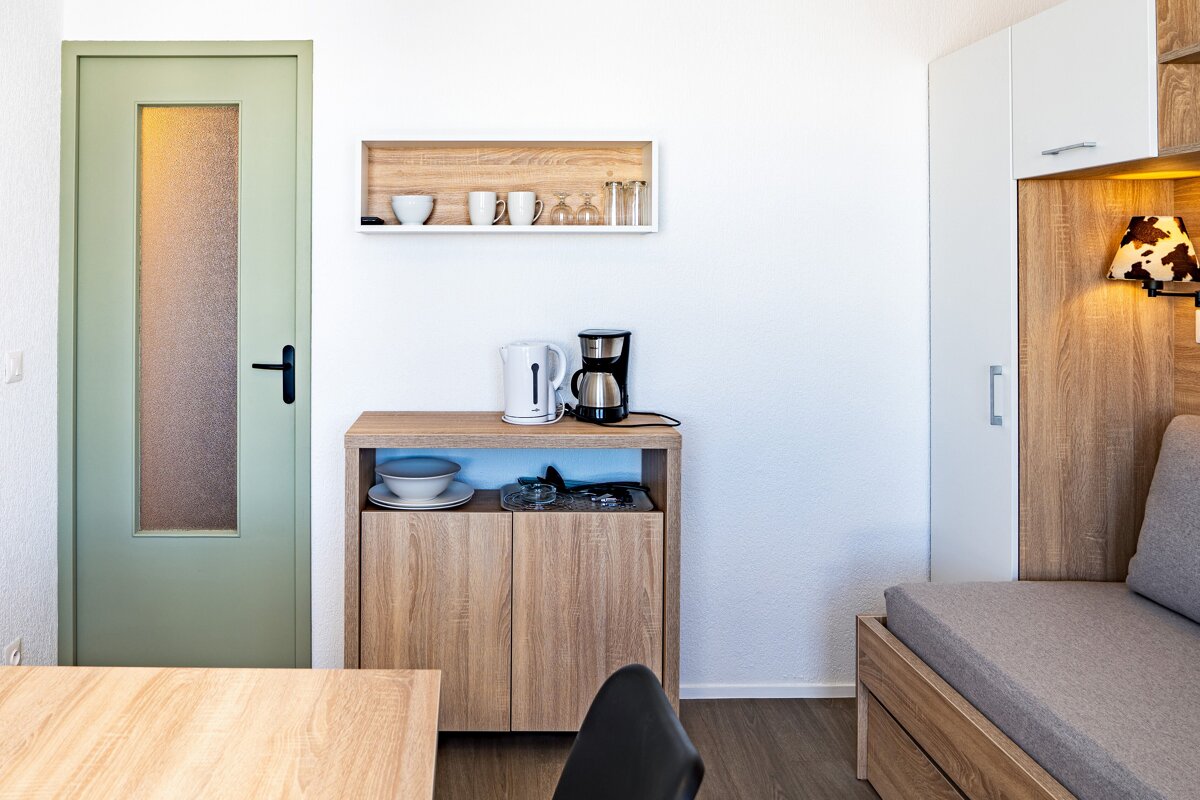 A bright, modern kitchenette and living area with a light green door, wooden cabinet with coffee maker/kettle, shelf with dishes, and a sofa bed.
