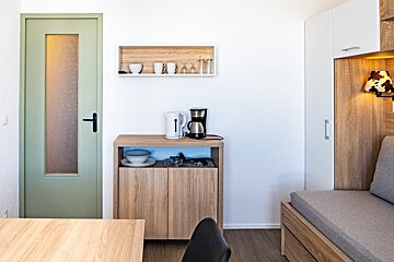 A bright, modern kitchenette and living area with a light green door, wooden cabinet with coffee maker/kettle, shelf with dishes, and a sofa bed.