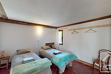 A simple bedroom with two single beds, white walls, exposed wooden beams, a window, and terracotta floor.