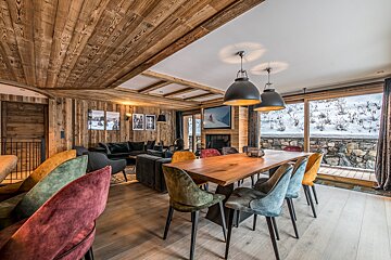 Alpine chalet with a wooden interior, modern dining area with colorful chairs, cozy living space, and large windows revealing a snowy view.