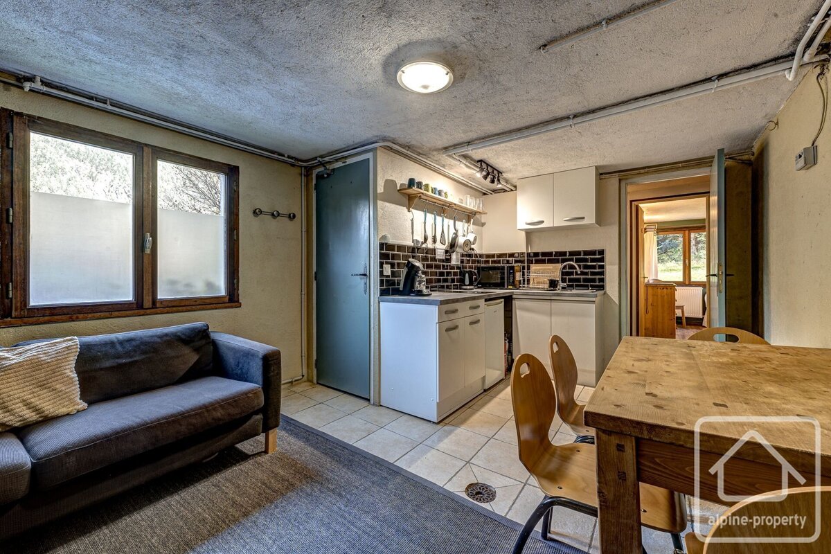 A compact, rustic studio apartment features a kitchen with white cabinets, a dining table, and a dark sofa. Exposed pipes line the ceiling, and a frosted window lets in light.