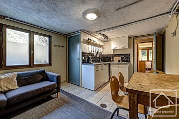 A compact, rustic studio apartment features a kitchen with white cabinets, a dining table, and a dark sofa. Exposed pipes line the ceiling, and a frosted window lets in light.