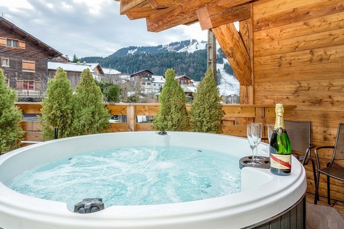 Bubbling hot tub with champagne on a wooden balcony overlooking snowy mountains and alpine chalets.