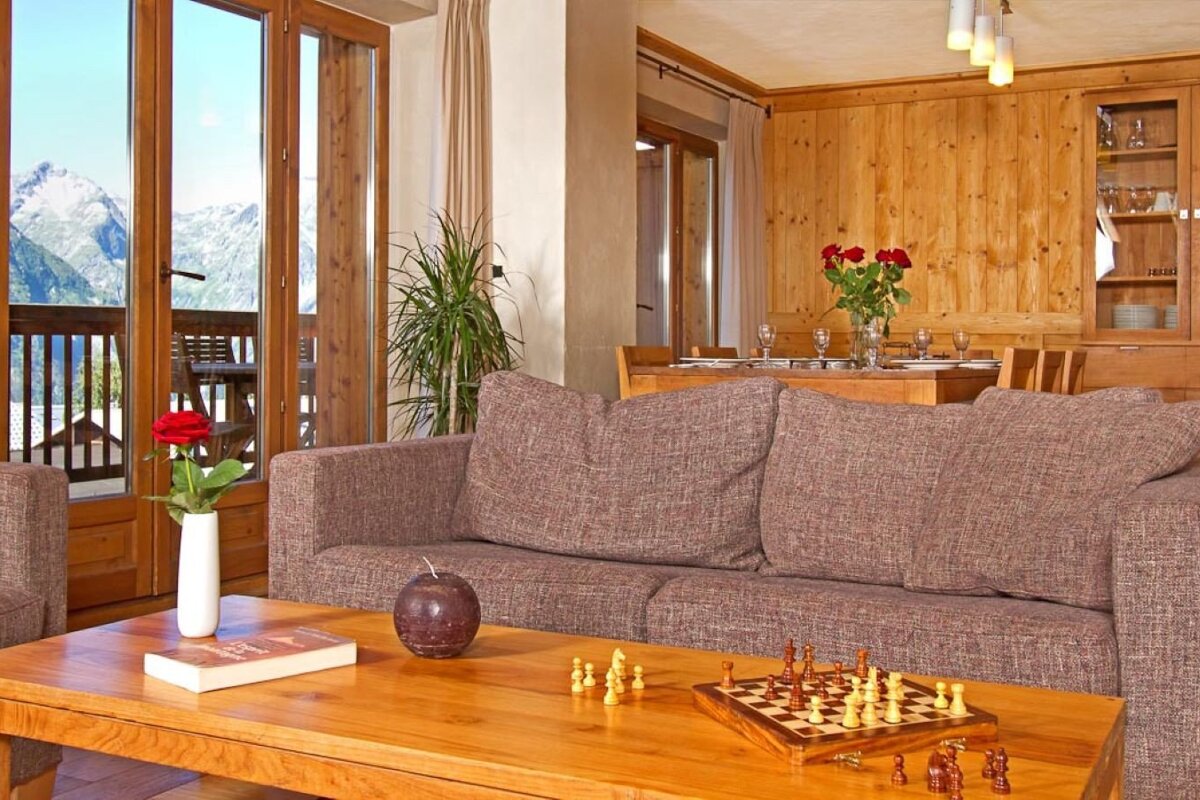 Cozy chalet living room with a sofa, chess set on a wooden table, and a balcony offering a stunning mountain view. Warm wood paneling throughout.