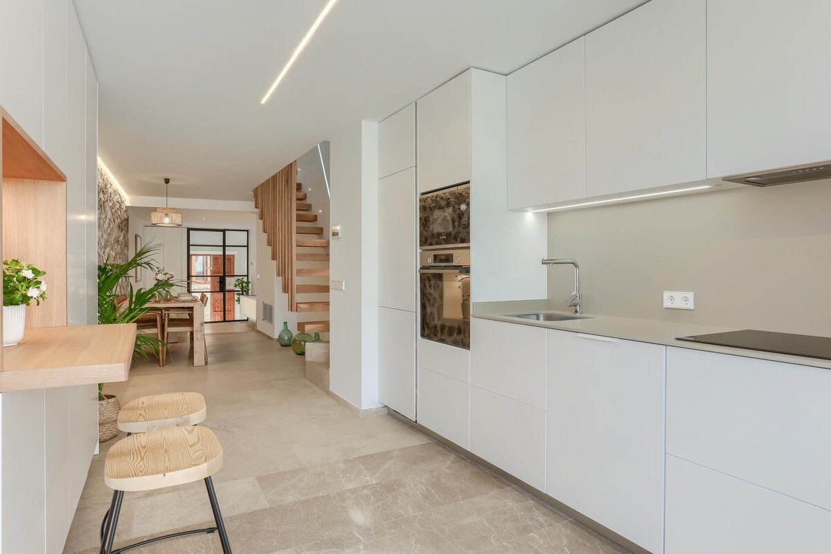 A modern open-plan home features a white minimalist kitchen with bar seating, leading to a dining area with a stone wall, and a wooden staircase.