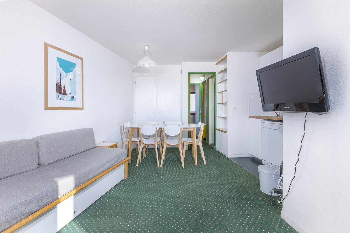 A bright living/dining area with a grey sofa, white dining table & chairs, green patterned carpet, wall-mounted TV, and a small kitchen visible.