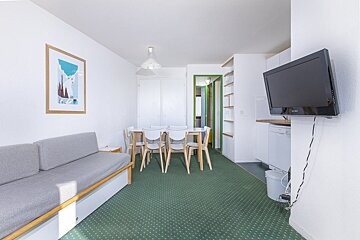 A bright living/dining area with a grey sofa, white dining table & chairs, green patterned carpet, wall-mounted TV, and a small kitchen visible.