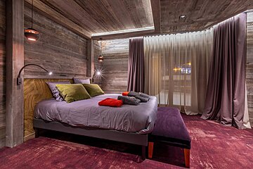 Cozy chalet-style bedroom with rustic wooden walls, a purple bed, olive pillows, and a deep red carpet. Warm lighting illuminates the space.