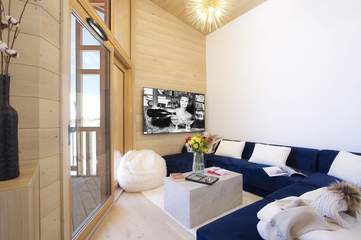 A cozy living room features wooden walls, a blue sectional sofa, a concrete coffee table, and a TV. A large glass door opens to an outdoor view.