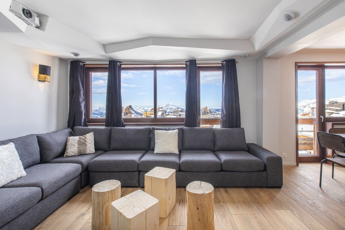 Cozy living room featuring a large grey sectional, wooden log tables, and windows overlooking scenic snowy mountains.