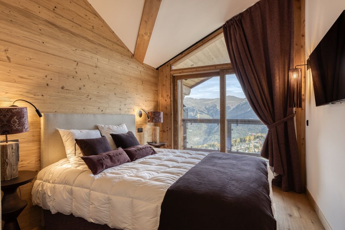 A cozy, modern bedroom with wooden walls, a plush bed, and a large window offering a stunning mountain and village view.