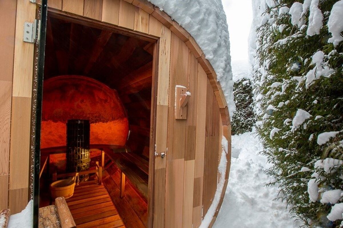 An outdoor barrel sauna with a glowing red interior stands in a snowy winter landscape next to snow-covered trees.