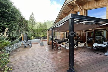 A rustic wooden deck features a covered outdoor lounge area, a hot tub, and a cozy chalet, all surrounded by lush greenery and mountains.