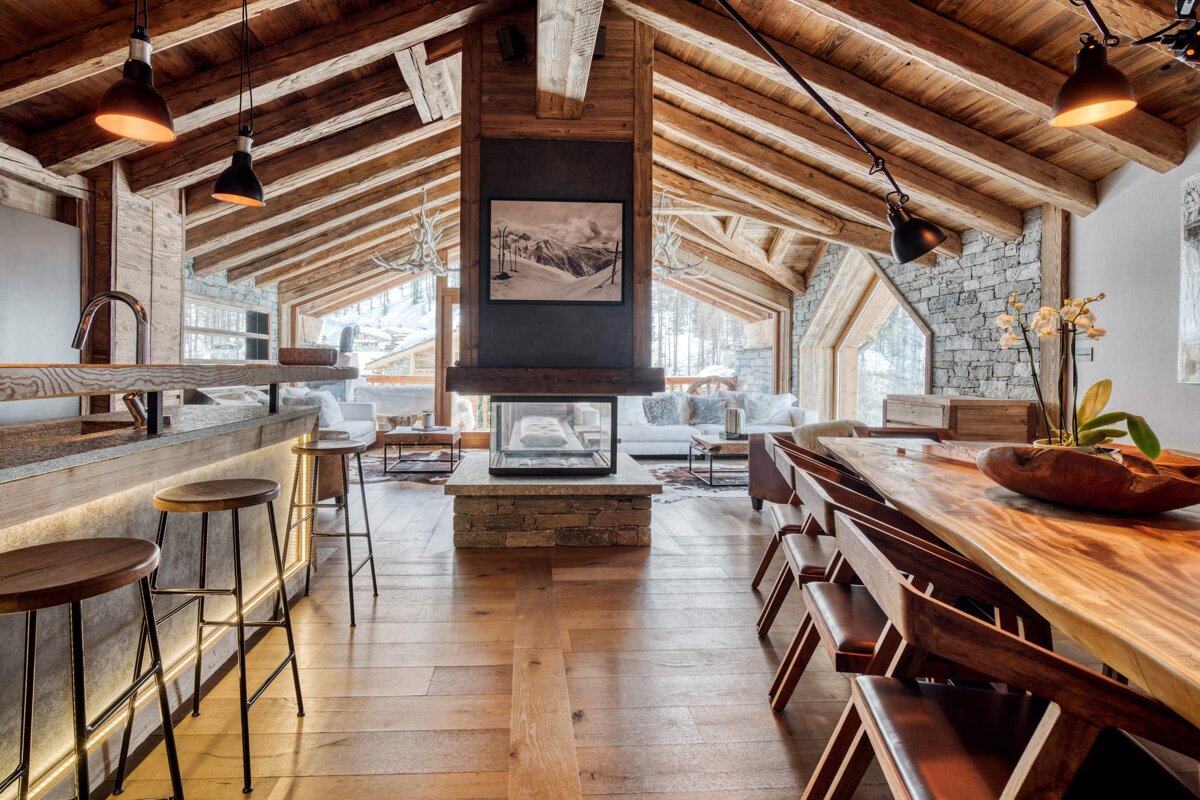 Luxurious modern chalet with vaulted wooden ceilings, open-plan kitchen, dining, and living areas centered around a fireplace, overlooking snowy mountains.