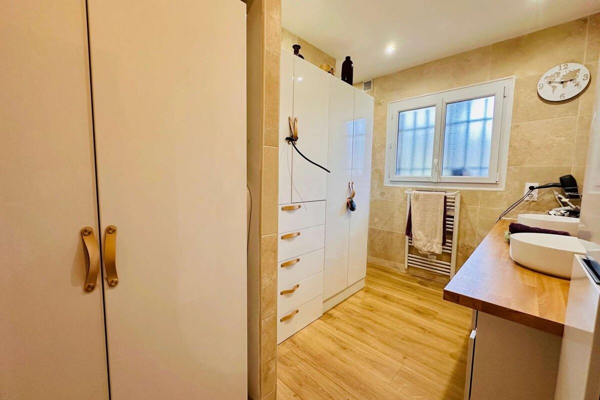 A bright bathroom features white cabinetry, wooden flooring, beige tiled walls, a window, and a double sink vanity.