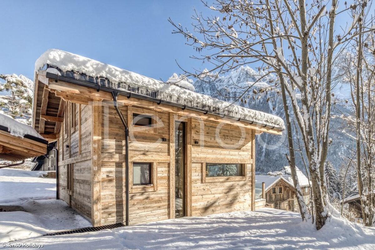 A rustic wooden chalet blanketed in snow and icicles stands amidst snowy trees, with majestic mountains under a clear blue sky in the background.
