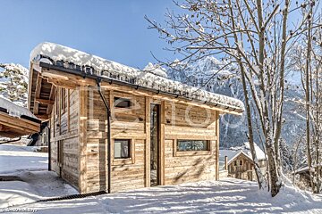 A rustic wooden chalet blanketed in snow and icicles stands amidst snowy trees, with majestic mountains under a clear blue sky in the background.