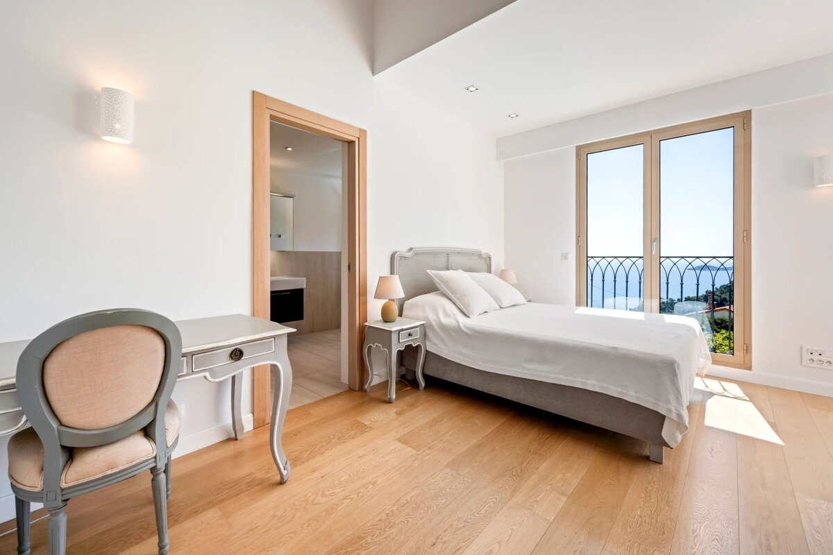 Bright, modern bedroom with wooden floors, grey bed and matching desk, featuring a balcony with sea views and a doorway to a bathroom.