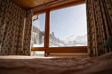 A cozy bedroom view through a large window, looking out at a bright, snow-covered mountain landscape.