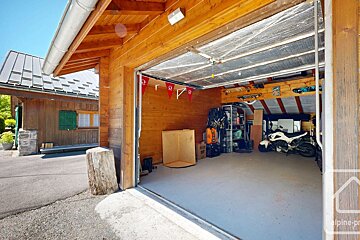 An open wooden garage with a motorcycle, storage, and red flags inside. Outside, a driveway leads to another wooden building with a gray roof under a clear sky.