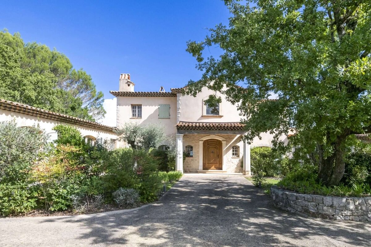 A beige Mediterranean-style villa with a long driveway, framed by mature trees and vibrant green bushes under a bright blue sky.
